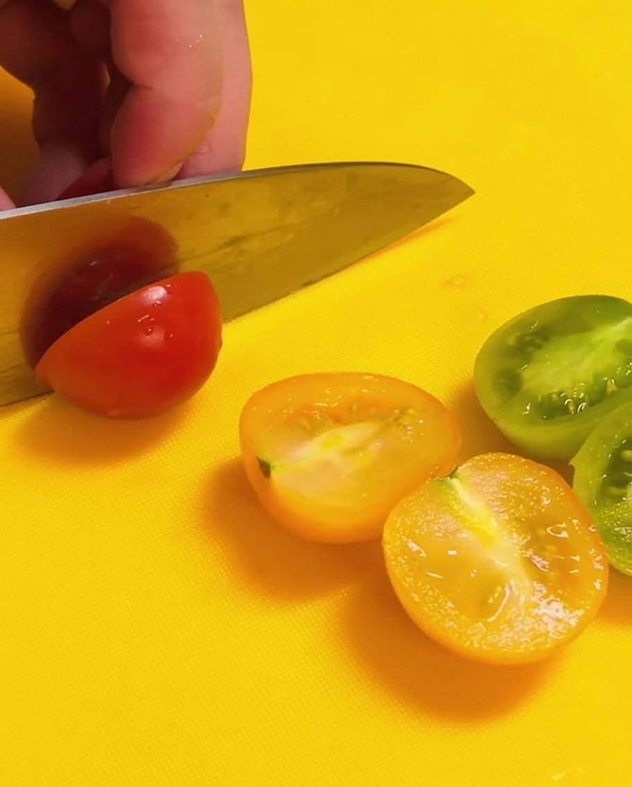 A knife slicing red and green cherry tomatoes in half on a bright yellow cutting board.