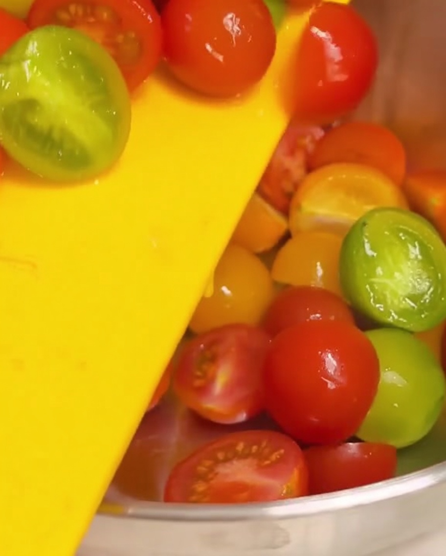 Halved cherry tomatoes being pushed off a yellow cutting board into a stainless steel mixing bowl.