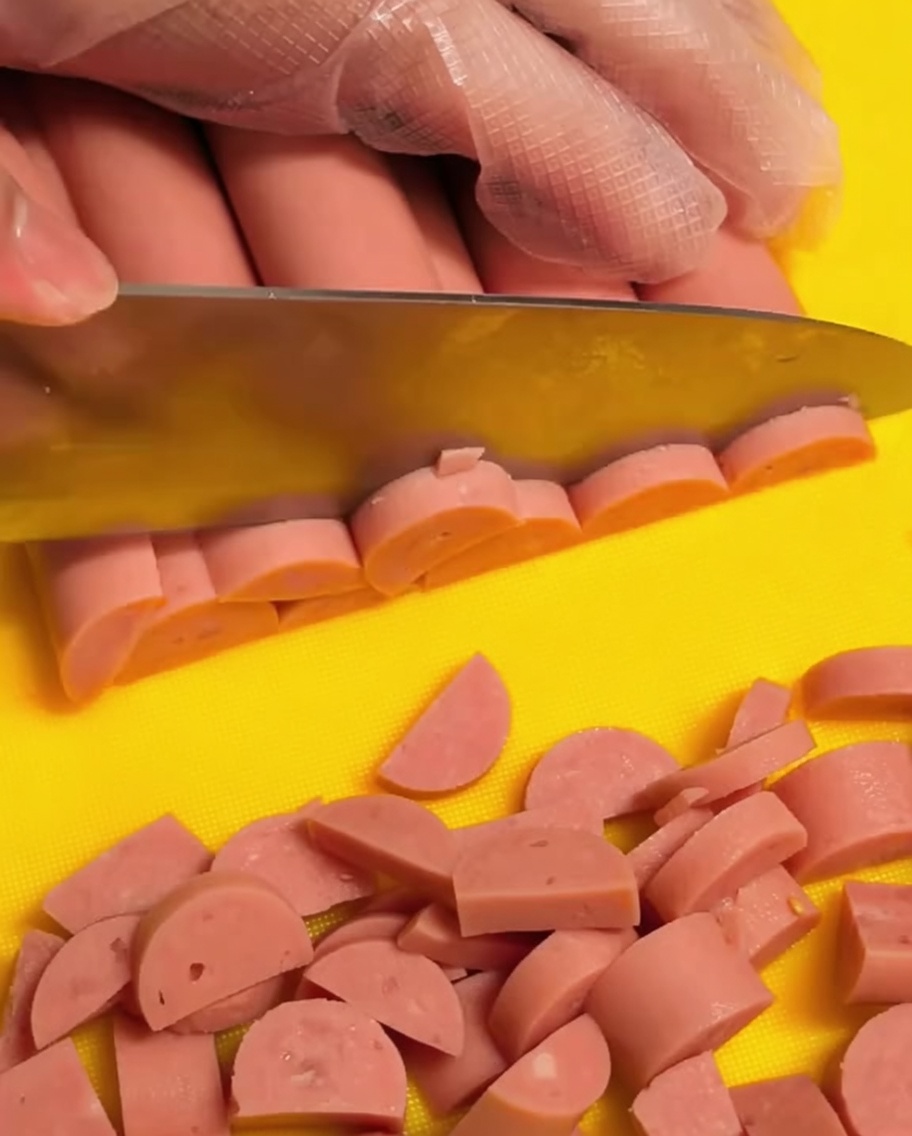 A hand wearing a clear glove slicing pink ham sausages into small half-moon shapes on a yellow cutting board.