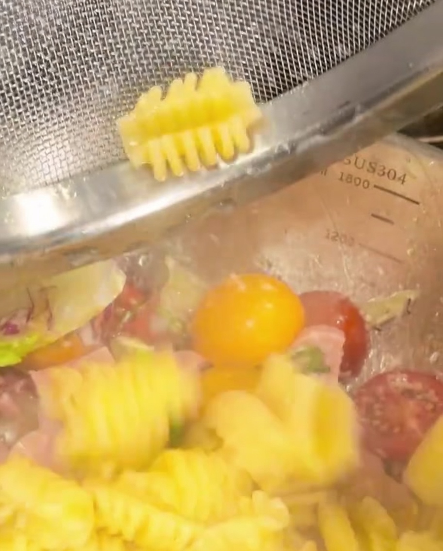 Cooked spiral pasta being transferred into a bowl with vegetables and ham.