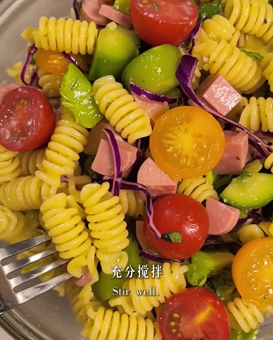 Pasta and salad ingredients being mixed together with olive oil in a bowl.