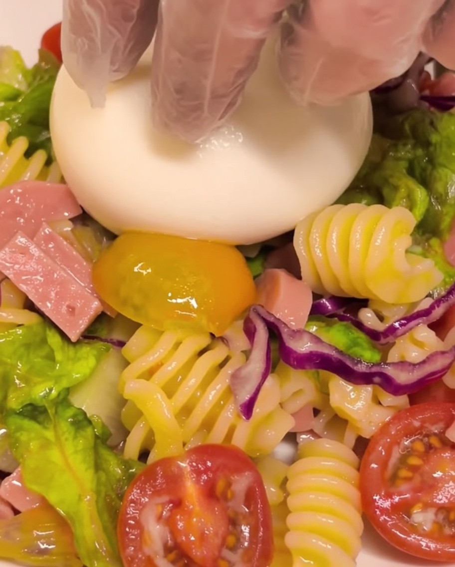 A fresh burrata cheese ball being placed in the center of a serving of cold pasta salad.