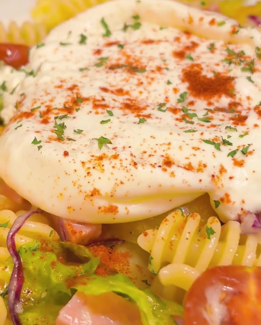 Close-up shot of the finished cold pasta dish featuring a creamy burrata center, garnished with parsley and red pepper, with a final golden drizzle of olive oil.