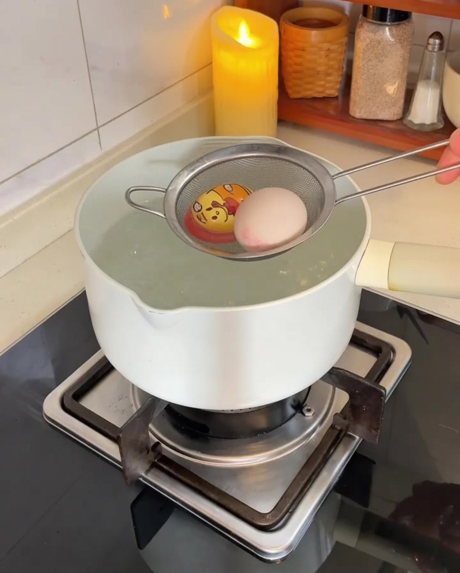 Eggs being carefully lowered into a pot of boiling water using a stainless steel mesh strainer.