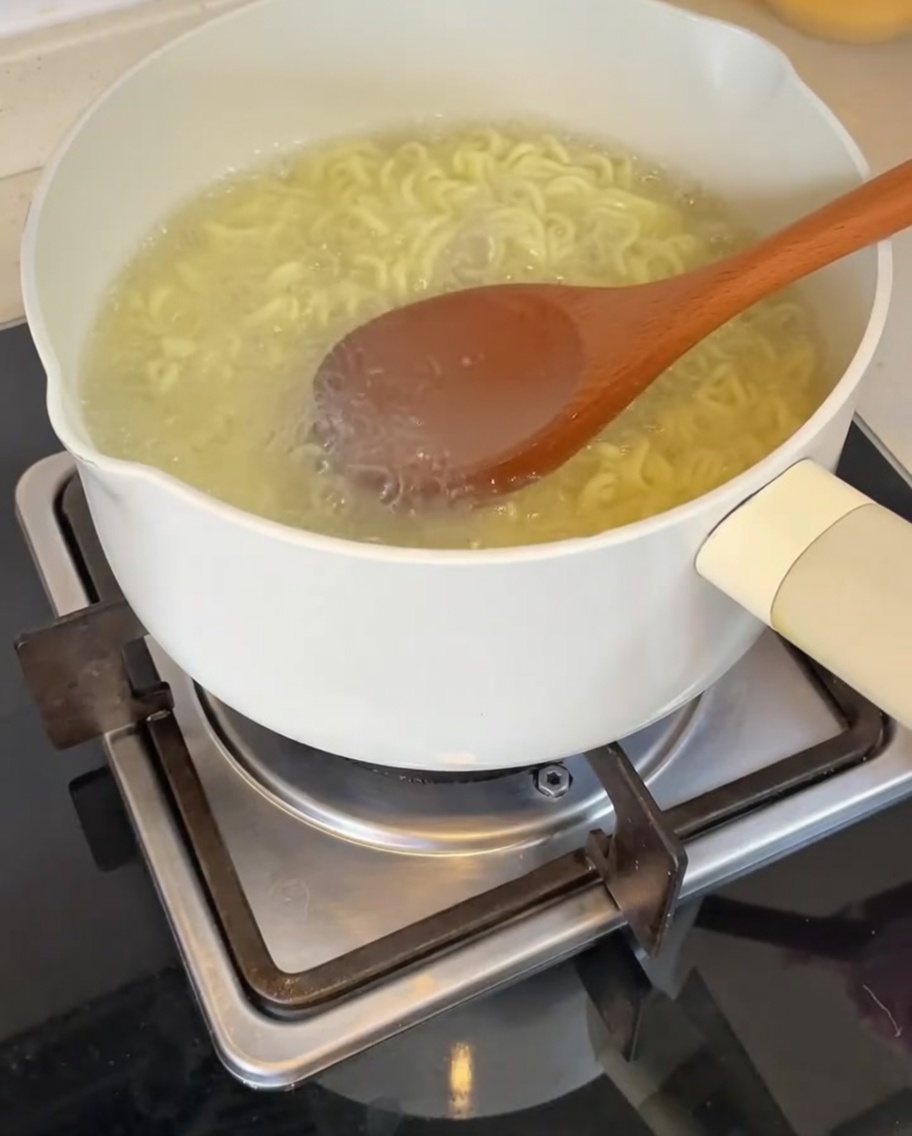 A wooden ladle pouring hot, cloudy noodle cooking water into the bowl containing the prepared sauce base.