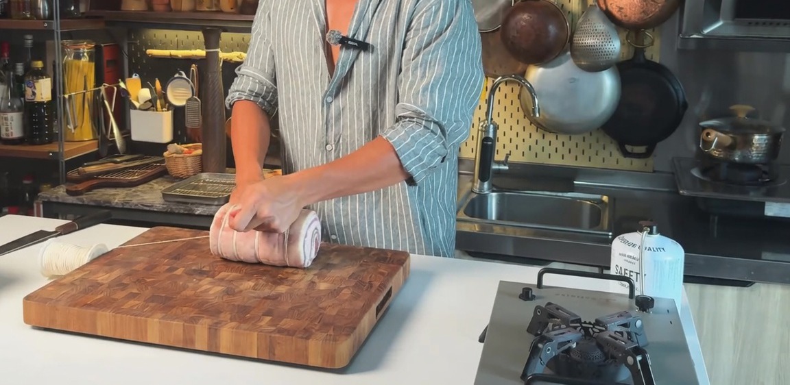 A rolled raw pork belly being tied horizontally with kitchen string on a wooden cutting board.