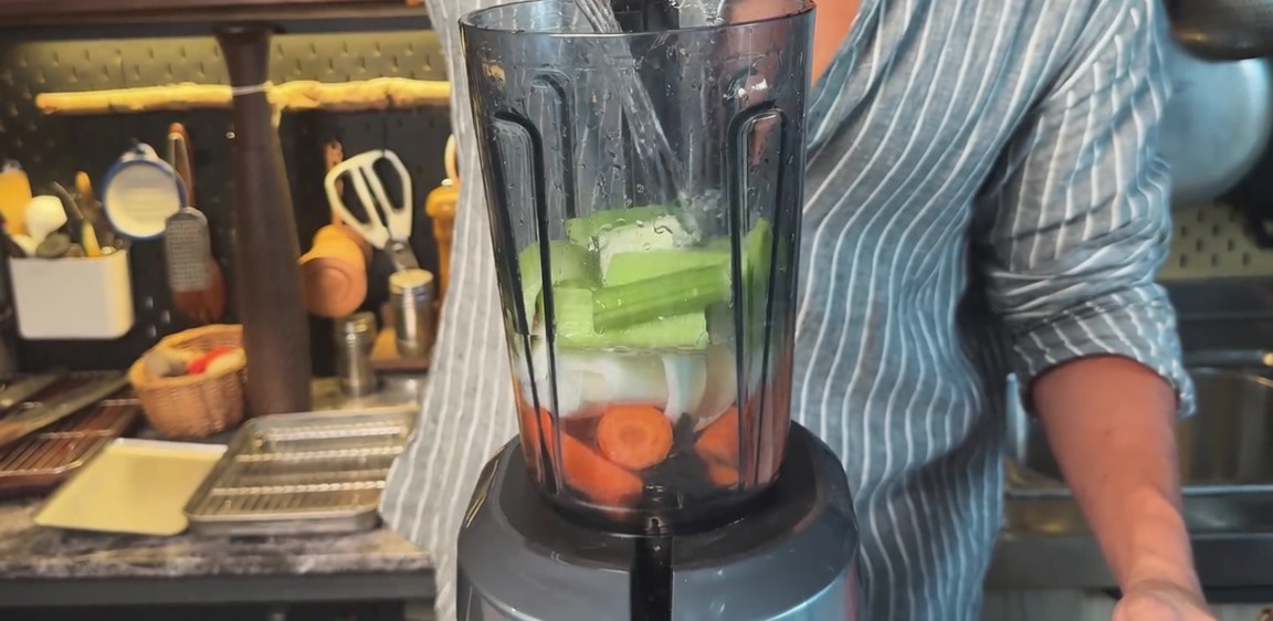 Pouring water into a blender filled with chopped carrots, onions, and celery.