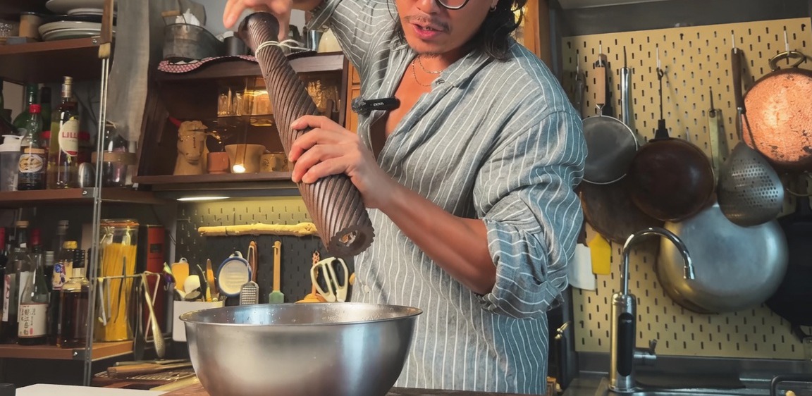 Using a tall wooden pepper grinder to season a marinated pork belly in a metal bowl.
