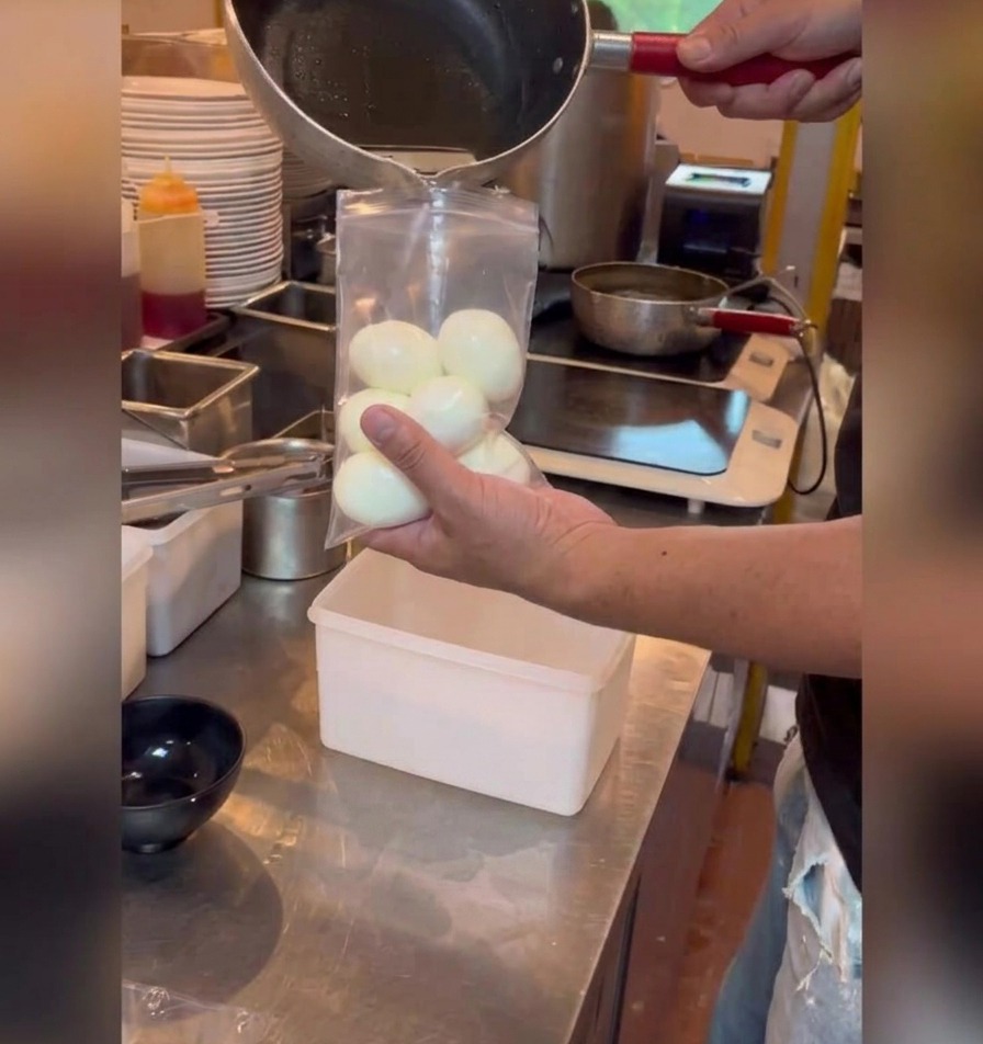 Pouring a dark soy-based marinade from a small saucepan into a clear plastic bag filled with peeled soft-boiled eggs.