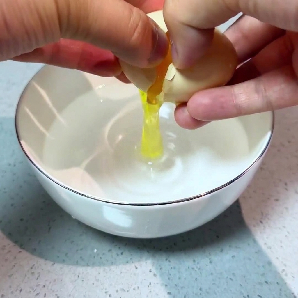 Two hands carefully cracking a fresh egg into a white bowl containing clear water.