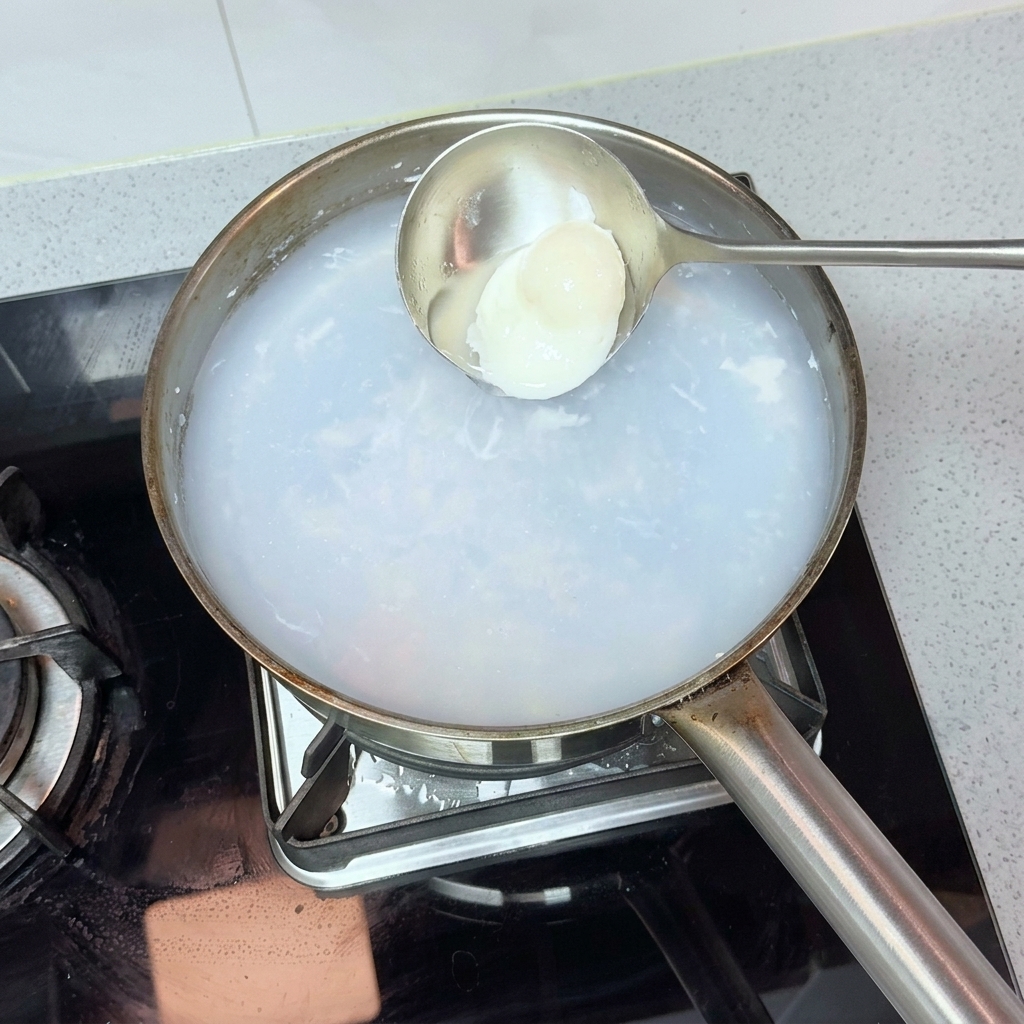 A metal ladle gently scooping a perfectly poached egg out of a pot of hot, cloudy water.