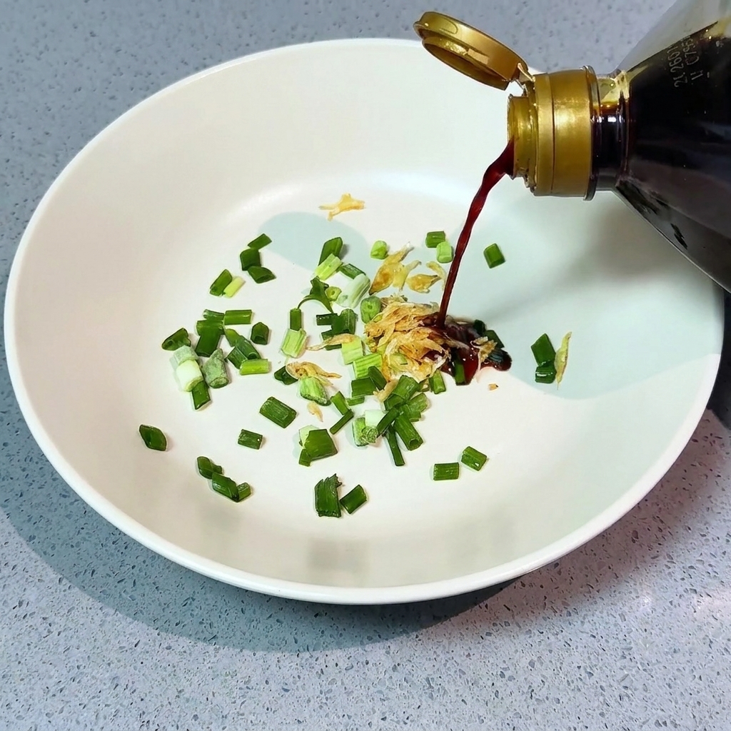 Pouring light soy sauce from a bottle over chopped green onions and dried shrimp in a white bowl.