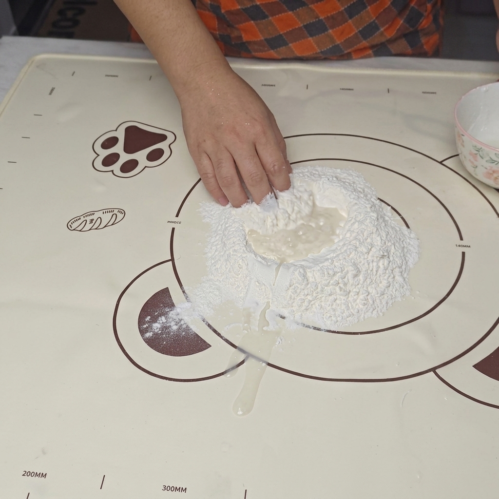 A hand mixing water into a well made in the center of a pile of white flour on a baking mat.