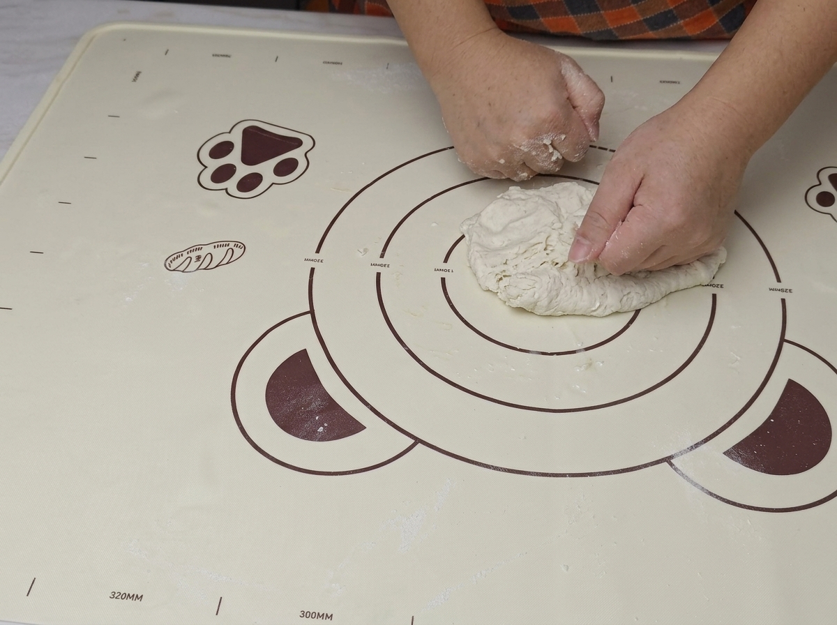 Two hands firmly pressing and folding a rough ball of dough on a silicone baking mat.