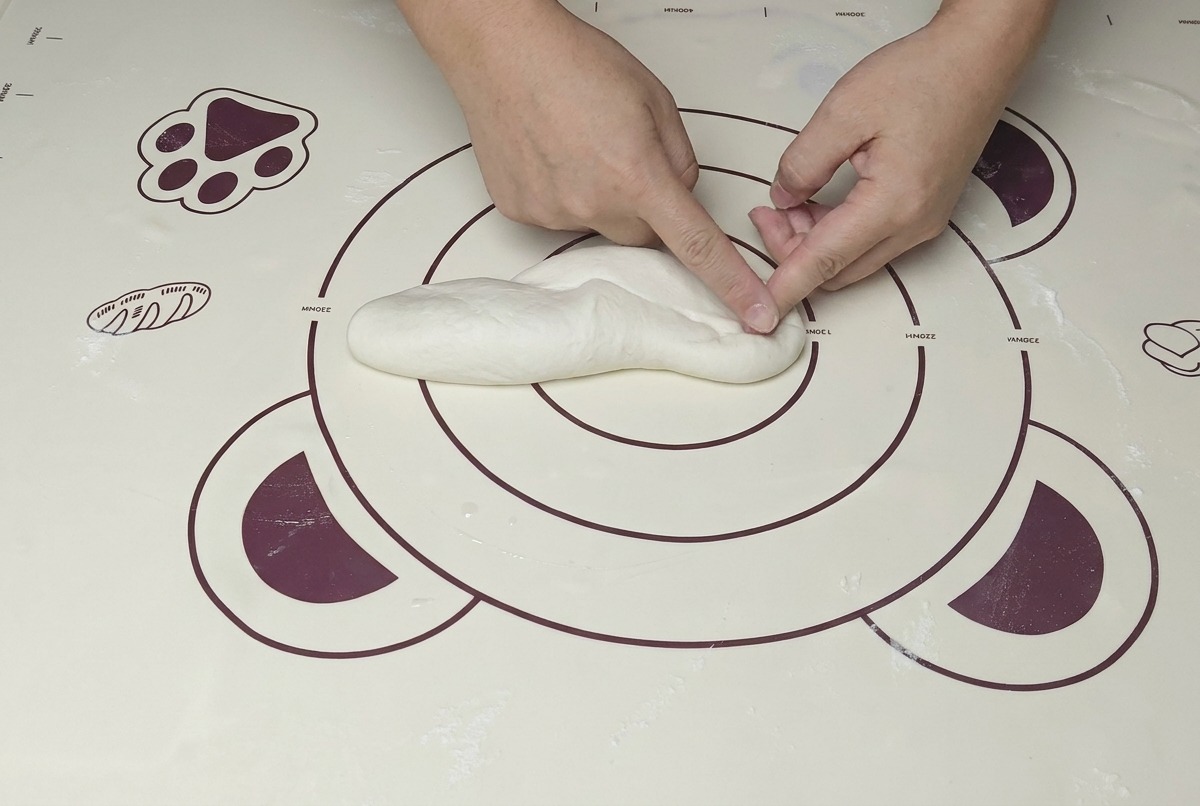 A pair of hands kneading a ball of dough on a mat, using the heel of the hand to press firmly.