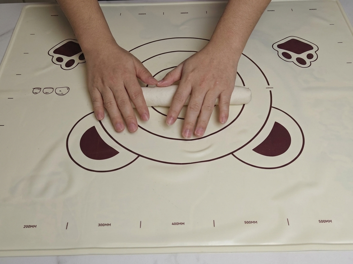 Two hands rolling a smooth piece of dough into a long, thick cylindrical log on a baking mat.