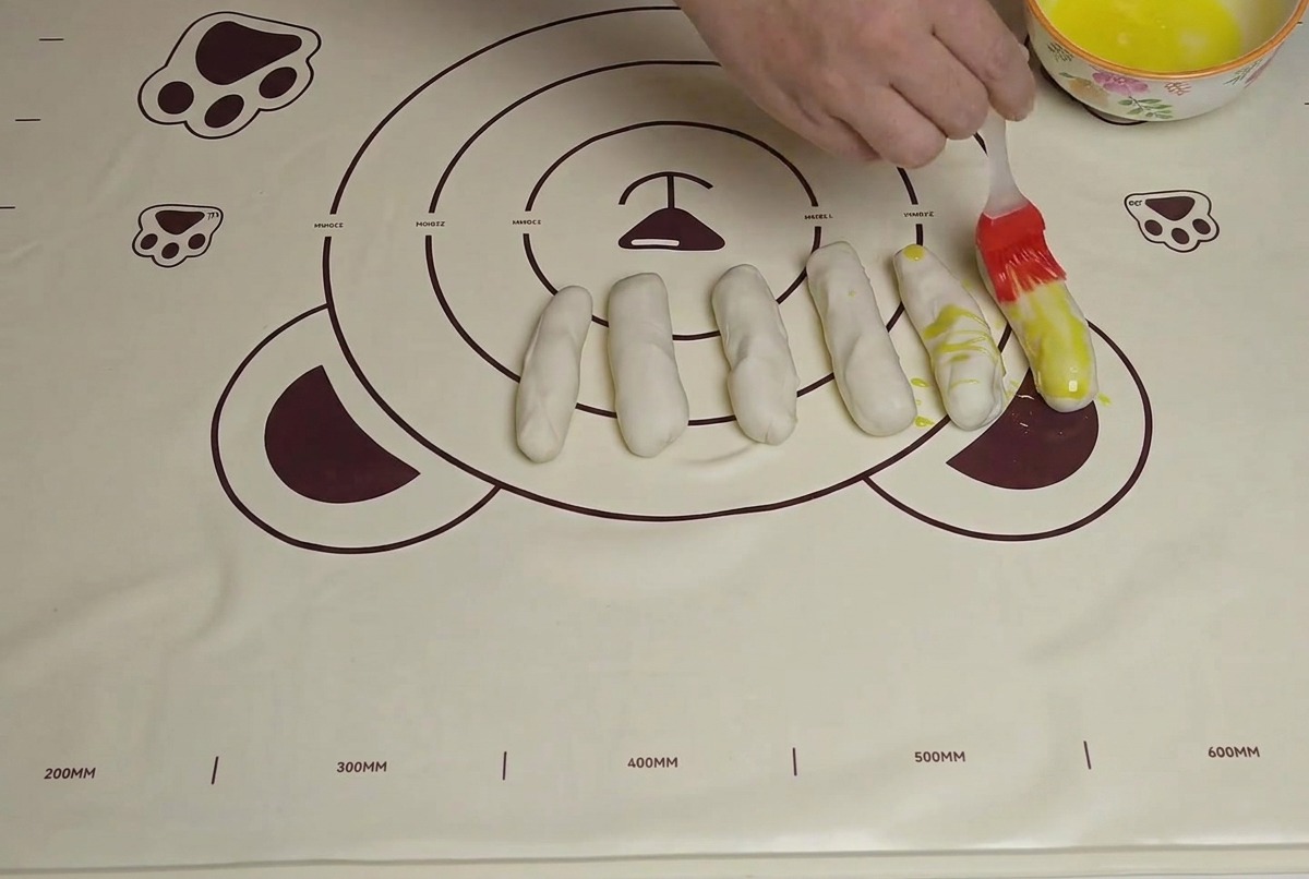 Multiple dough strips arranged on a mat, being brushed with yellow oil using a silicone brush.