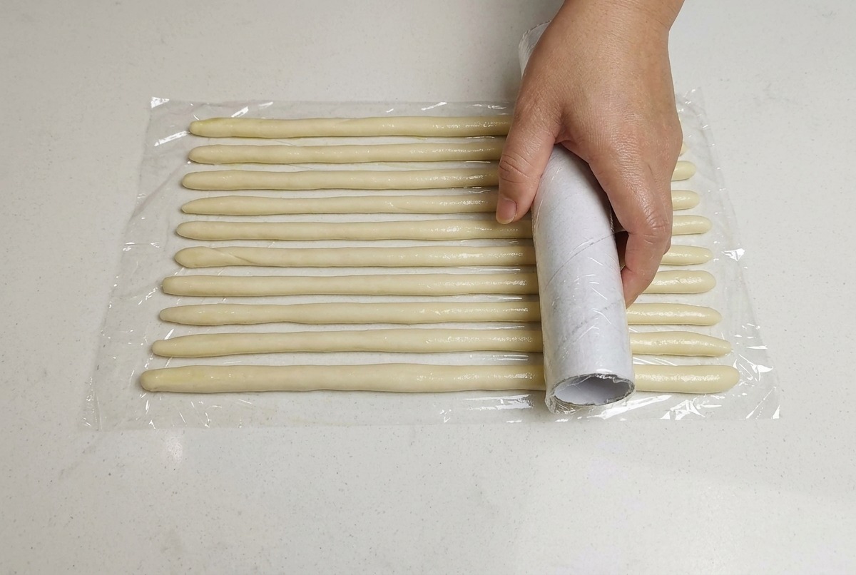 A hand placing a sheet of plastic wrap over arranged thin dough ropes on a white surface.