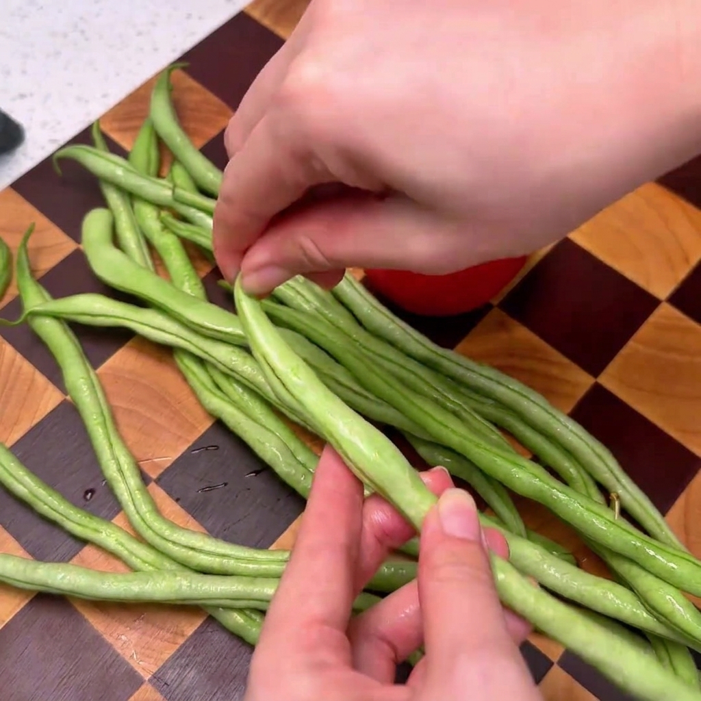 Green beans being trimmed on a checkered wooden cutting board.