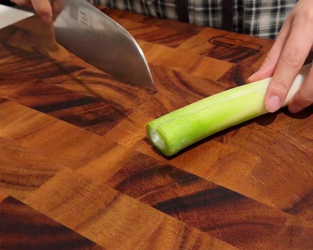 Hands carefully slicing a green onion into thin rings on a dark wooden cutting board.