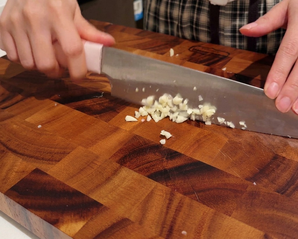 Hands holding a knife, mincing garlic cloves into small, fine pieces on a wooden cutting board.
