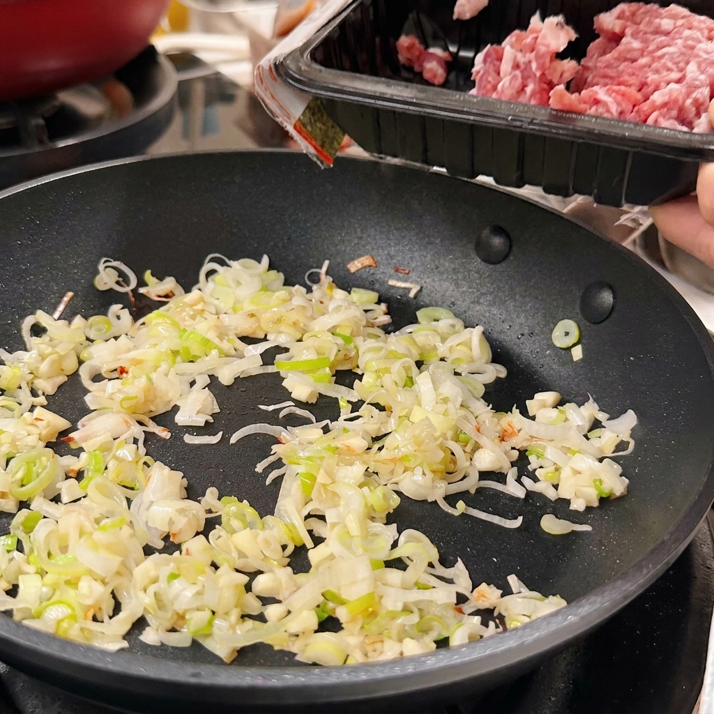 Raw minced pork being slid from a black plastic tray into a pan with sautéed green onions and garlic.