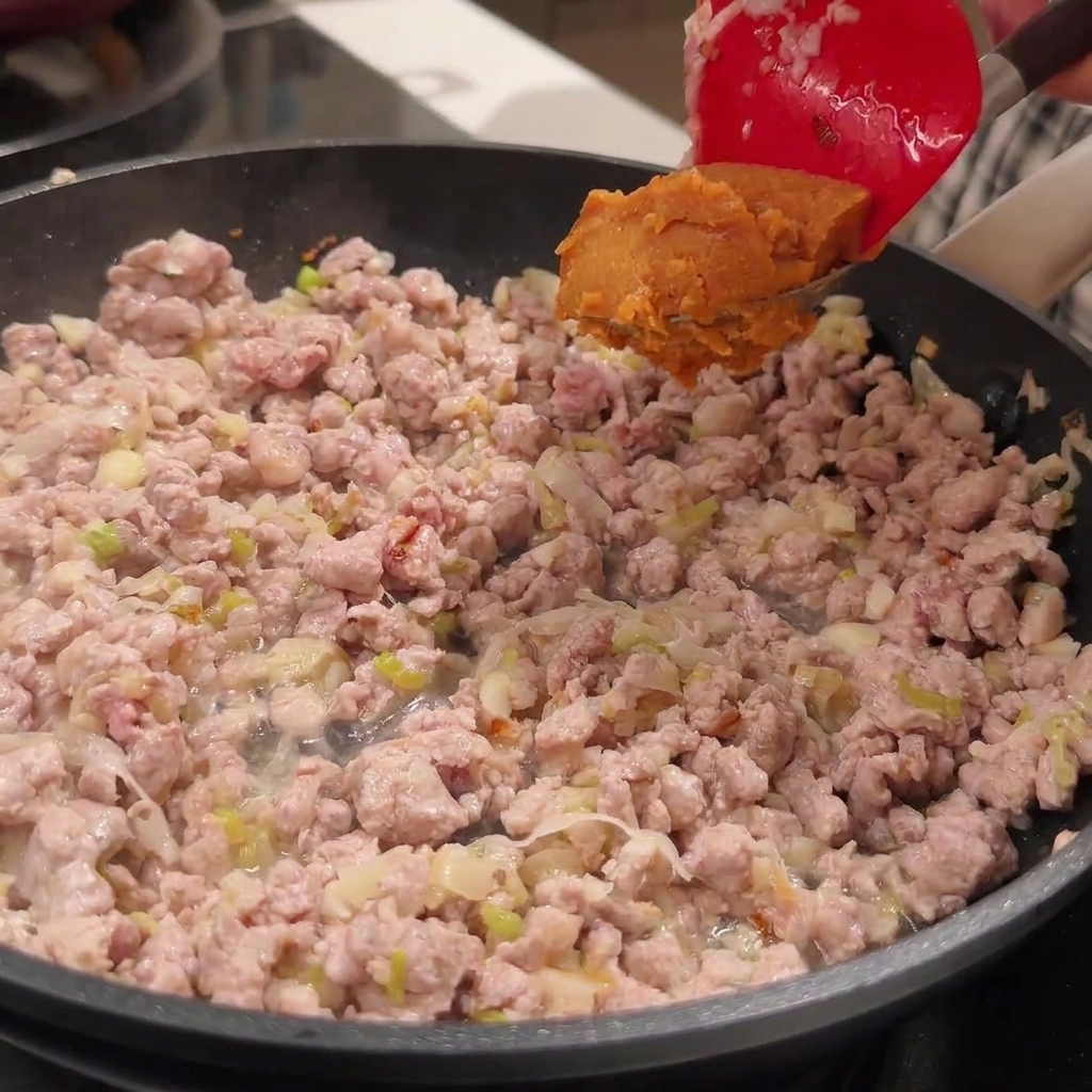 A red spatula holding a dollop of miso paste above a pan filled with browned minced pork.