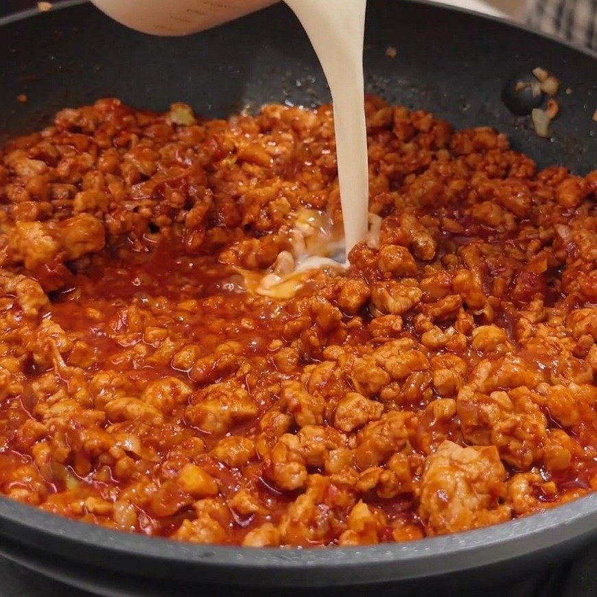 Creamy white soy milk being poured from a clear glass measuring cup into a pan of simmering, rich red miso meat sauce.