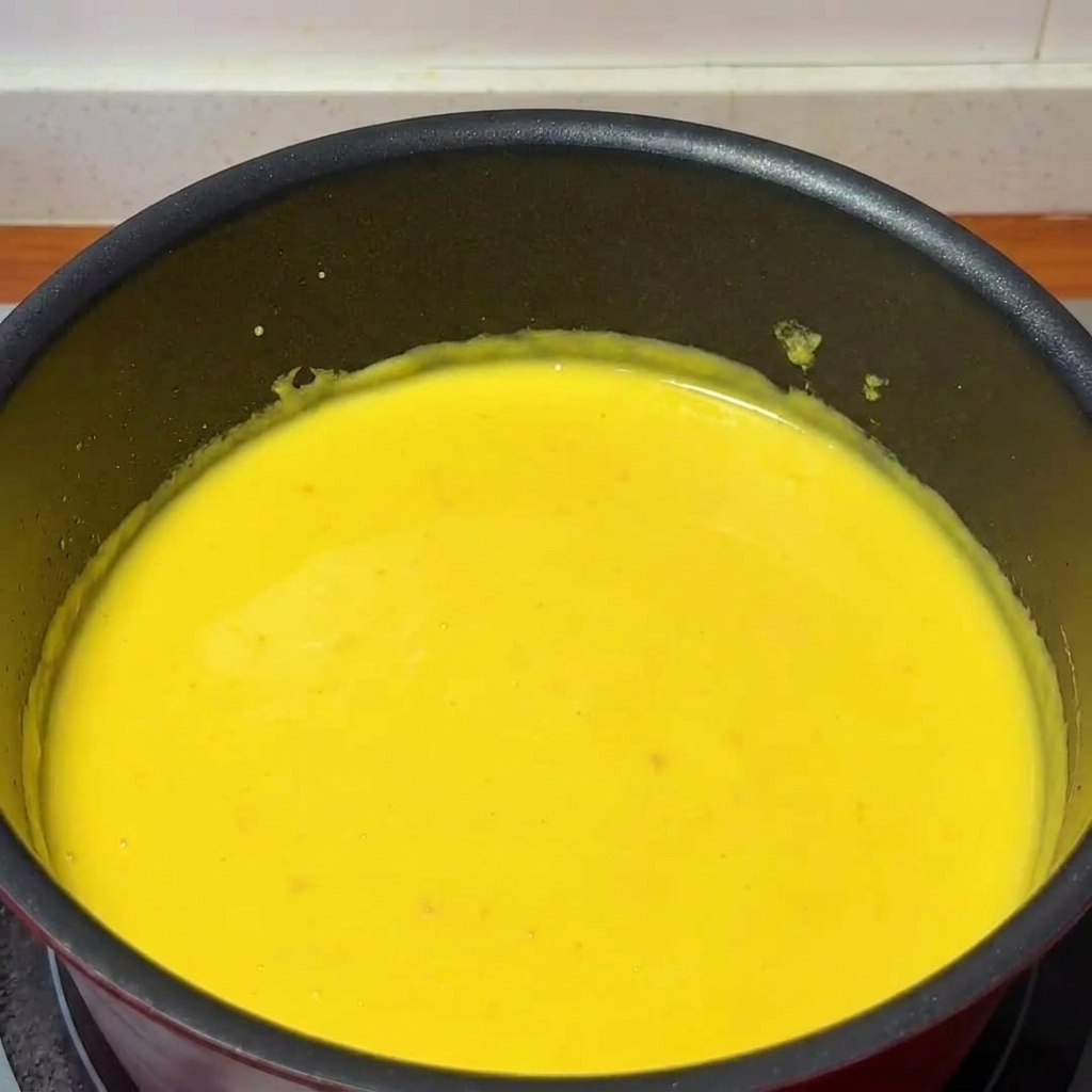 A ladle pouring fresh yuzu juice into a saucepan filled with thick, creamy cheese sauce, being stirred by a wooden spoon.