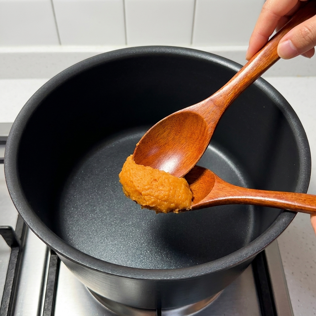 A wooden spoon stirring miso paste, mirin, sugar, pork bone broth, and yuzu juice in a saucepan over a stove.