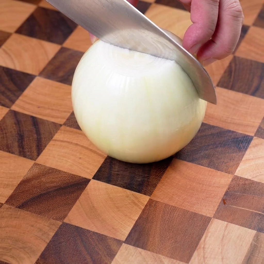 Yellow onion being carefully sliced into thin strips on a wooden cutting board.