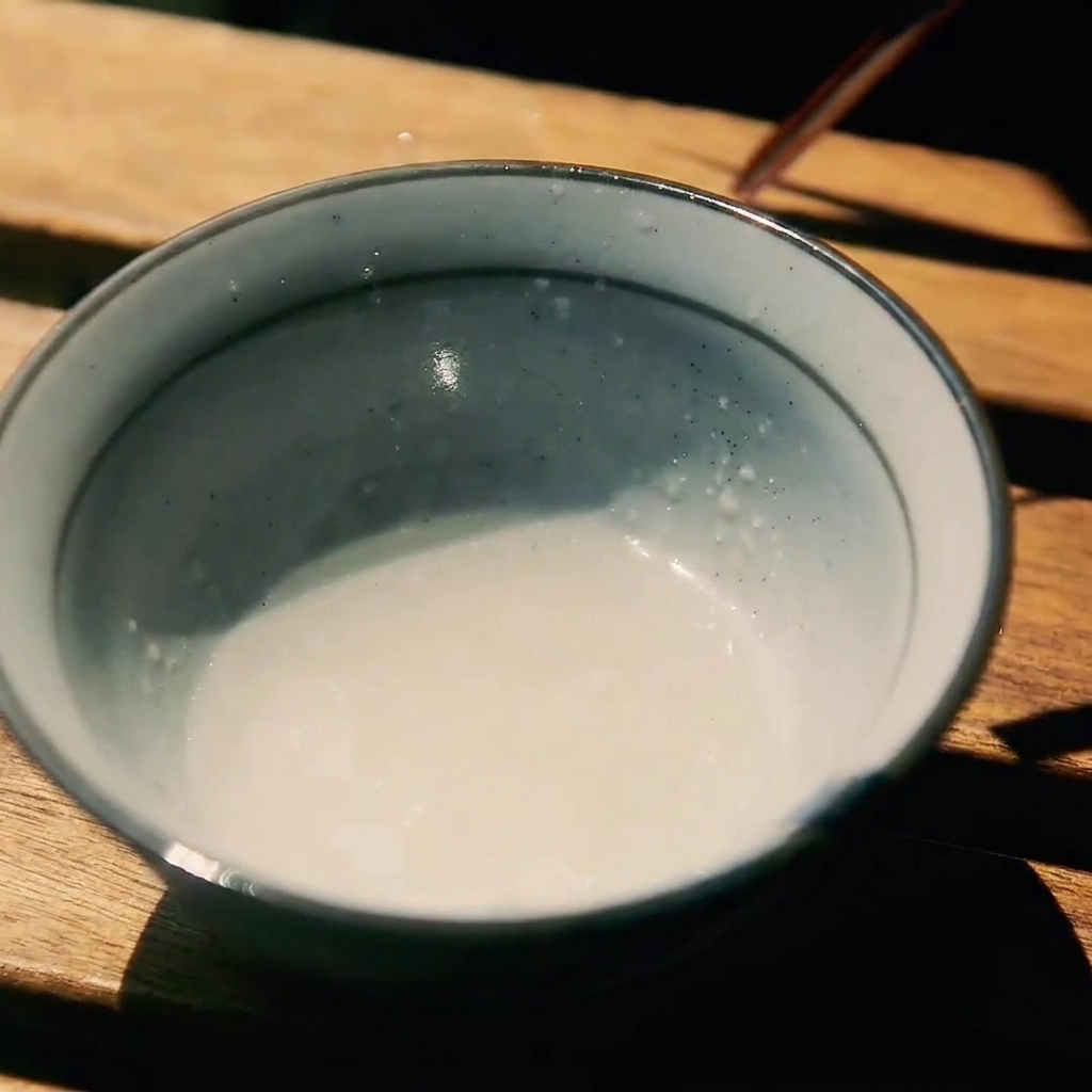 A small ceramic bowl on a wooden surface containing a mixture of sake, sugar, and soy sauce.