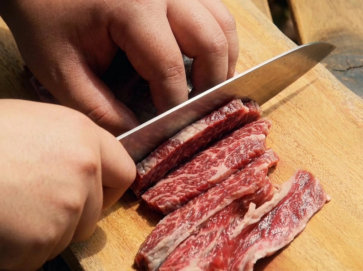 A sharp knife slicing raw beef short ribs into thin strips on a wooden cutting board.
