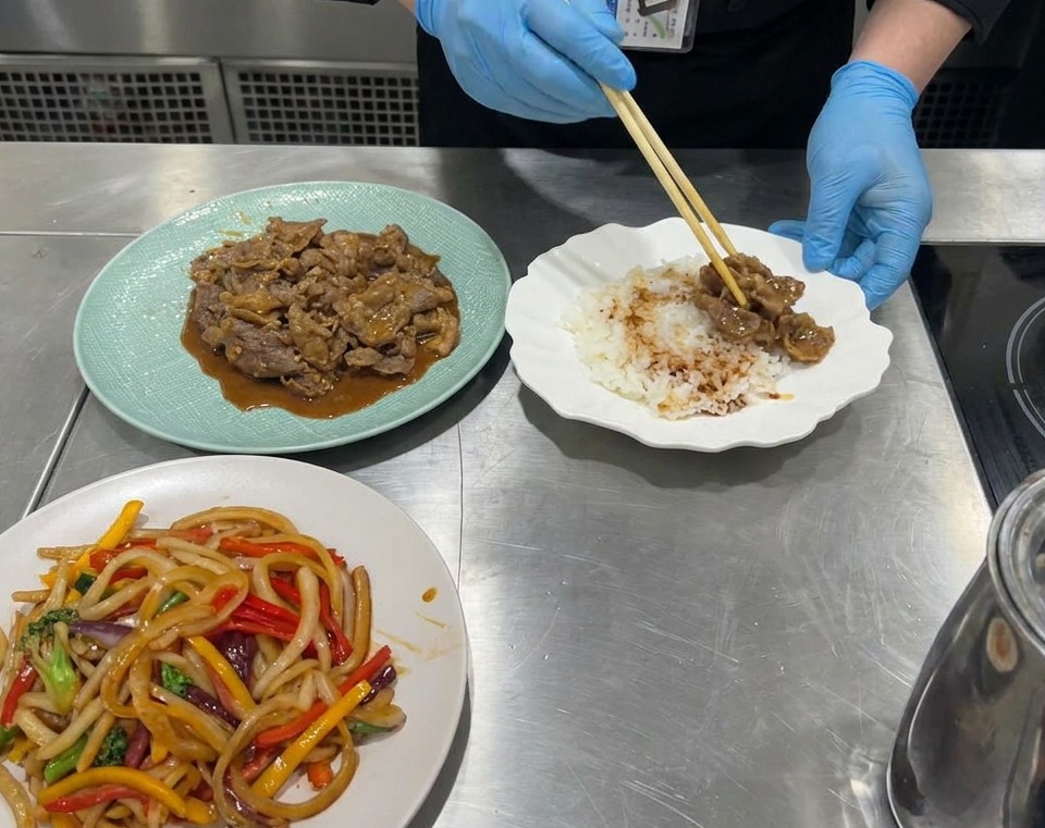 A close-up view of a serving bowl being filled with a base of white rice, followed by portions of cooked beef and colorful stir-fried vegetables.