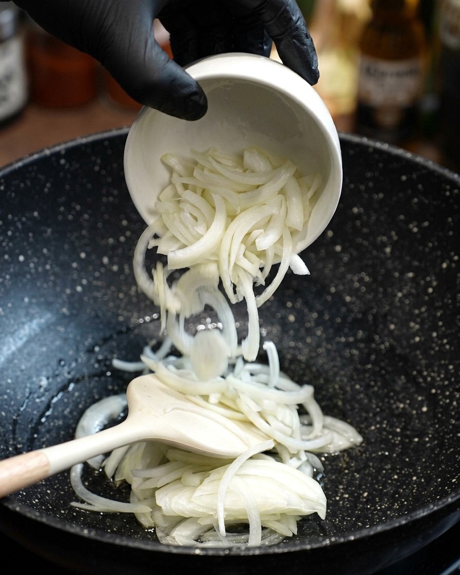 Freshly sliced white onions being added to a pan with hot oil and stirred with a spatula.