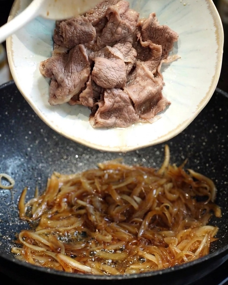 A dark, savory teriyaki sauce being poured into a pan with sautéed onions and beef.