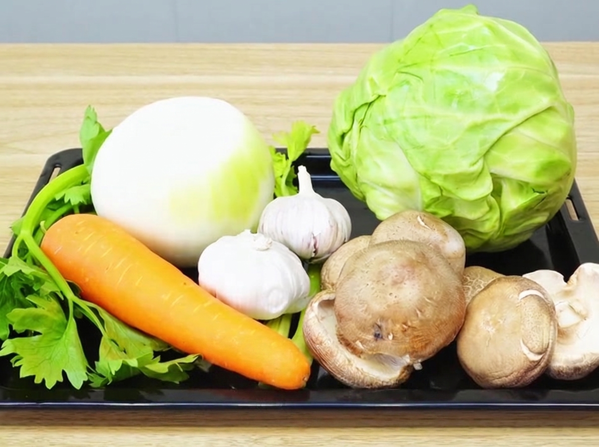 A variety of fresh vegetables including onion, garlic, carrot, celery, shiitake, and cabbage neatly arranged on a wooden cutting board.