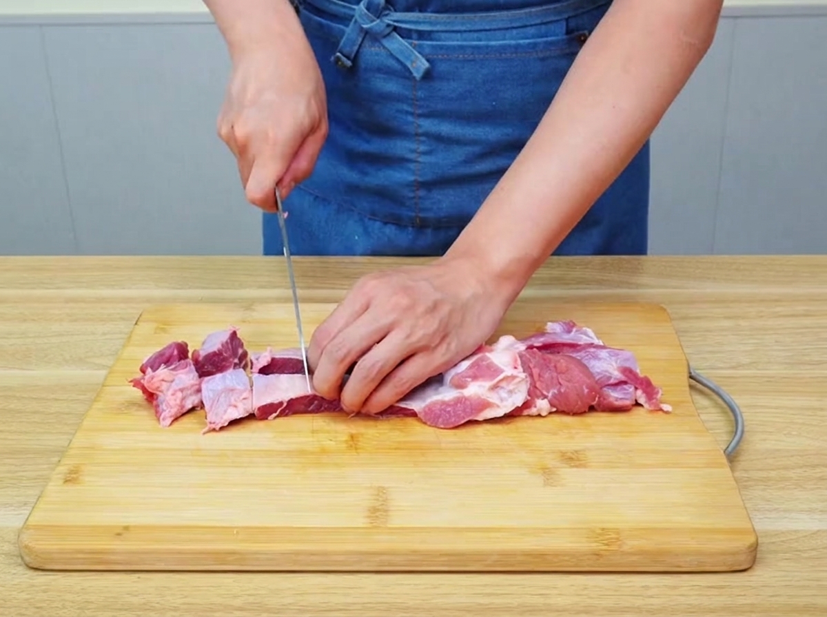 Raw beef brisket being carefully cut into large, uniform chunks on a wooden cutting board.