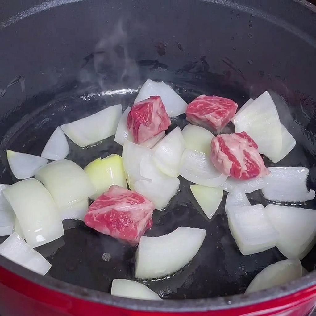 Beef cubes cooking alongside onions in a red pot.