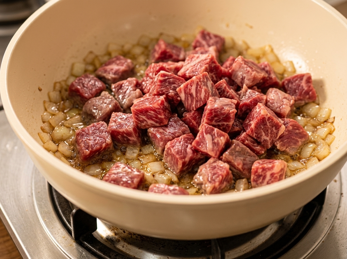 Cubed beef being added to a pan containing sautéed onions and diced vegetables.