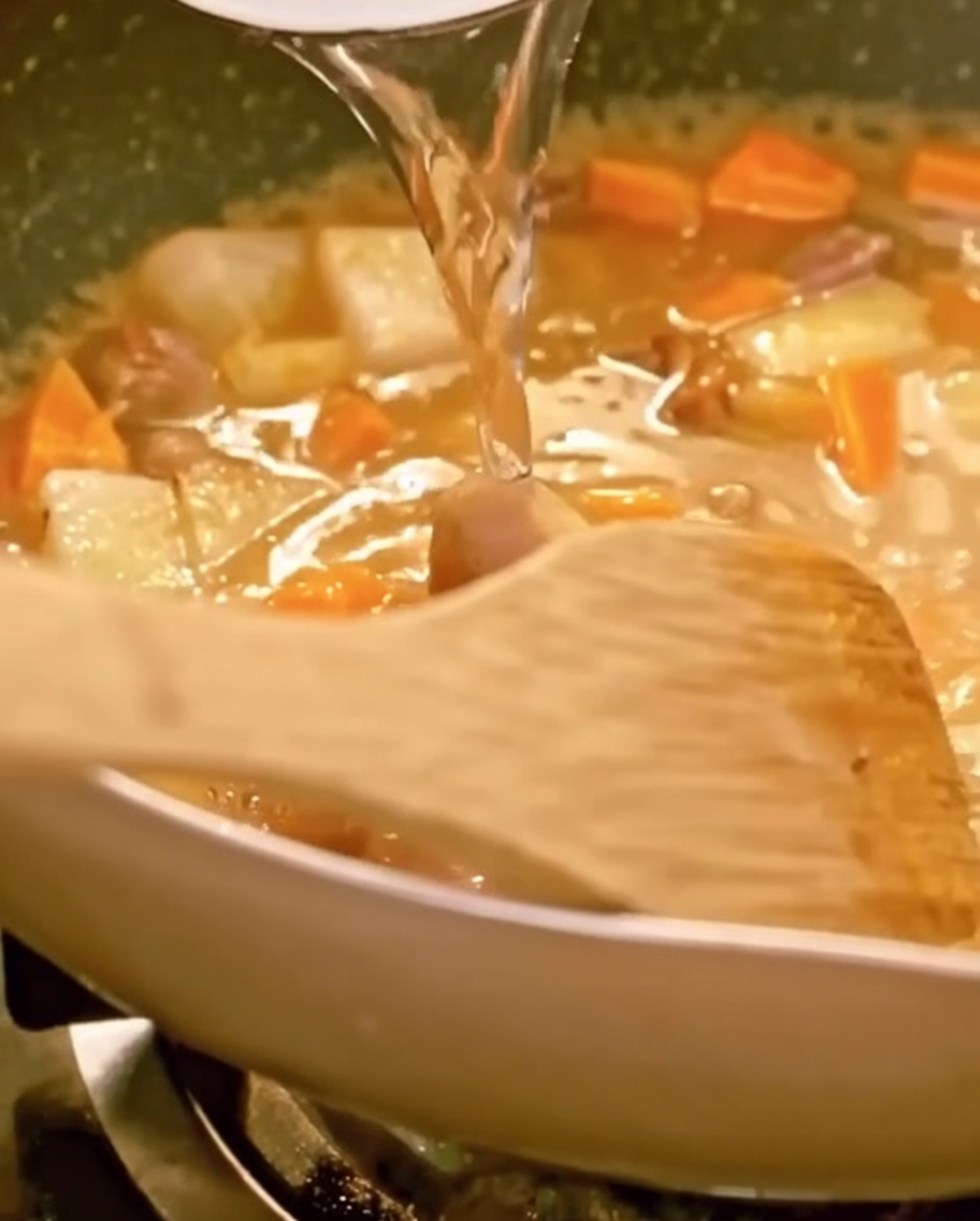 Water being poured into a pan filled with beef, vegetables, and melting curry, with a wooden spatula visible.