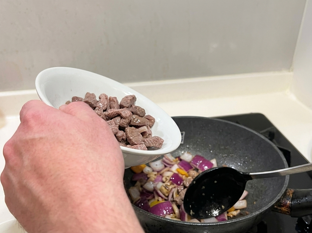 Blanched beef chunks being mixed with sautéed red onions in a pan.