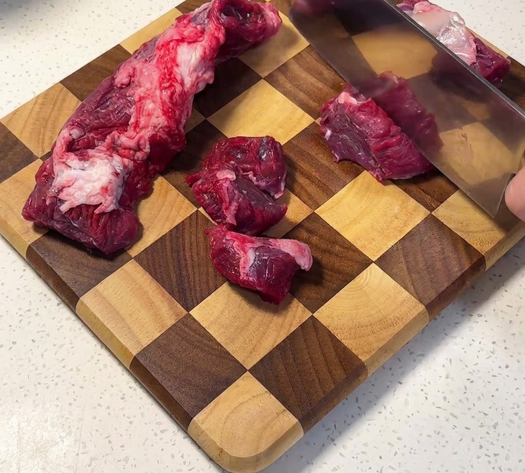 Beef brisket being cut into uniform cubes on a checkered wooden cutting board.