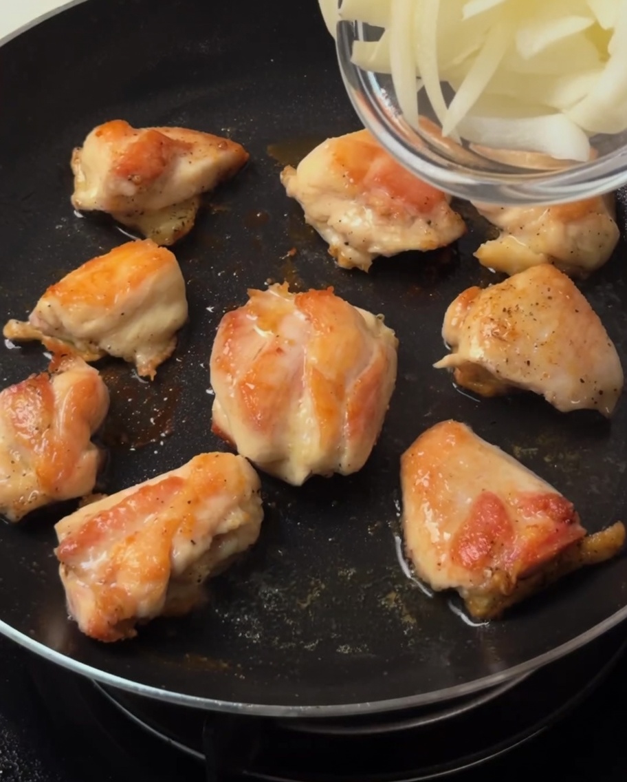 Sliced onions being stirred into the pan with the browned chicken pieces.