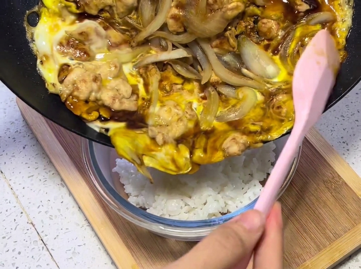 A close-up of the chicken, egg, and onion mixture being transferred from the pan onto a serving bowl of steaming white rice.