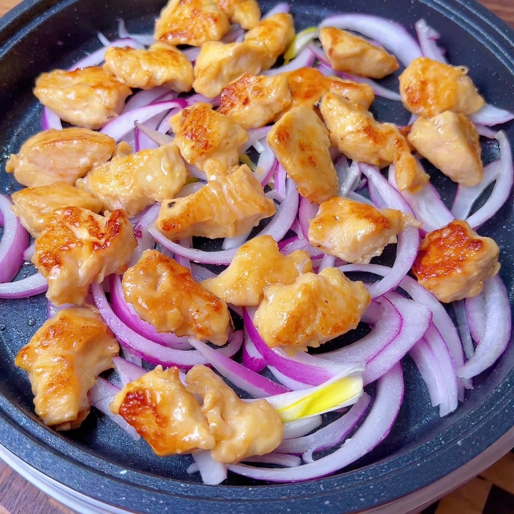 Golden brown chicken pieces and translucent onion slices being coated with a savory sauce in a pan.