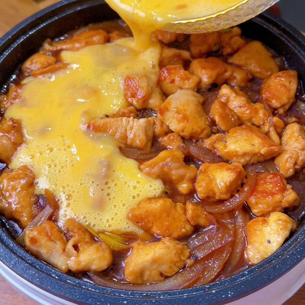 Beaten eggs being poured over the cooked chicken and onion mixture in a pan.