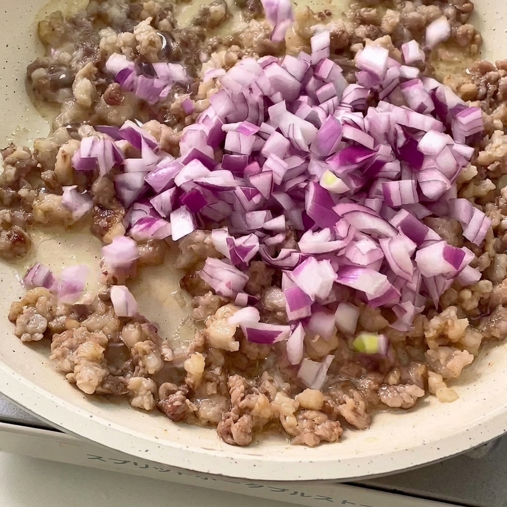 Chopped red onion and minced garlic being stir-fried with browned meat in a pan.