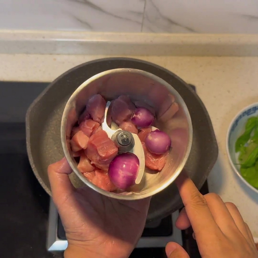 Manual meat grinder containing chunks of meat and red onion.