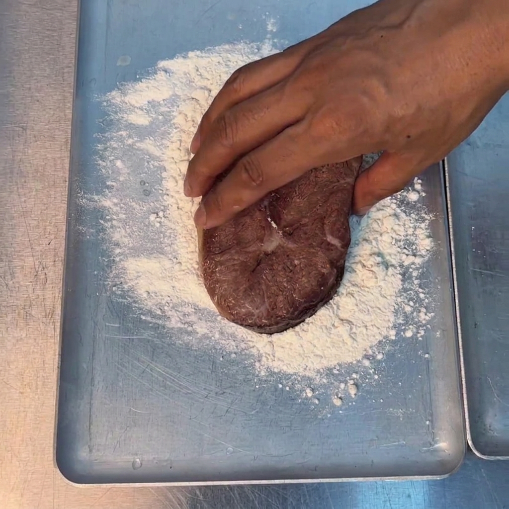 A hand pressing a thick cut of meat into white flour on a metal baking tray.